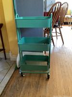 Front view of a green 3-tier metal trolley cart with wheels on a hardwood floor near kitchen and dining area furniture.
