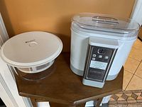 White electric yogurt maker device with a control panel on front and clear container with white lid on the left side of table