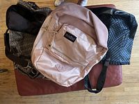 Top view showing all three bags: Jansport rose gold knapsack on top, brown patterned overnight bag underneath, and black woven tote bag to the side.