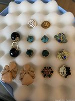 Top view of 9 pairs of Kate Spade earrings arranged on white foam. Shows various styles including black drop earrings, white studs, blue and green gemstone studs, bee designs, beige dangles, floral and signature face stud.