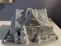 Front view of the house-shaped bundt cake pan showing detailed roof shingles, windows, door, and two trees in front.