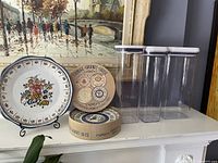 View of the three tall clear Oxo containers with white lids alongside two round Oxo cheese plates in their original boxes and the hand painted signed ceramic plate displayed on stand