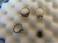 Top-down view of three rings and a pair of snowflake stud earrings on a white textured background. Two rings have clear heart-shaped stones in silver and rose gold, one ring has a rose gold leaf design, earrings have blue stones.