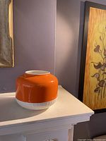 Angled side view of the orange and white ceramic vase sitting on a mantel against a purple wall with a framed painting to the right.