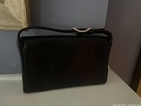 Front view of the black leather clutch showing smooth exterior, wrist strap with metal detail.