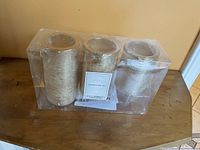 Three gold electric pillar candles in original clear plastic packaging on wood surface.