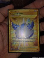 Full view of the gold holo Glitter full art Rugged Helmet Trainer card from Pokemon showing the card details, artwork, and condition.
