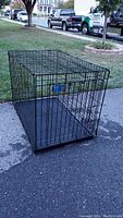 Black metal wire dog kennel on driveway, showing full side view with plastic tray inside.