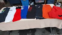 Seven men's short sleeve t-shirts and one pair of red shorts arranged on a table. Brands include Adidas, Reebok, Nike in various colors including white, black, blue, orange and red.