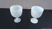 Two white milk glass goblets on black surface, displaying distinct textured patterns and scalloped rims