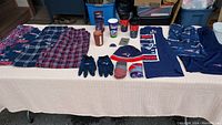 Wide view of all items laid out on beige tablecloth showing apparel and accessories including pants, shorts, gloves, beanie, cups, wallet, and candleholder.