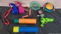 Top-down view of assorted water toys including water squirters, series of water blaster canisters, a green water gun, and several colorful ball toys.