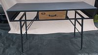 Front view of steel desk frame showing single wooden drawer inserted as a prop (not included). The desk frame is black with angled legs and support bars.