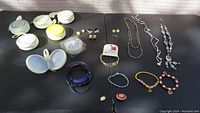 Photo showing multiple jewelry items laid on flat surface: white storage containers, earrings, necklaces, bracelets.