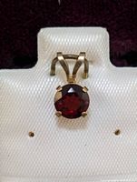 Front-facing view of the yellow gold pendant showcasing the round garnet stone in a six-prong setting, displayed on white fabric with '10 KT GOLD' text visible under the pendant.