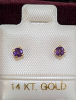 Close-up photo of the pair of 14K gold amethyst stud earrings displayed on white textured backing with '14 KT. GOLD' label.