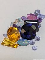 Photo showing assorted mixed genuine gemstones in various cuts and colors, including yellow citrine, deep blue stone, pink, purple amethyst, and opal stones, some with visible damage or breakage.