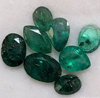 Photo of eight loose emerald gemstones in various shapes and sizes on a light background showing color and inclusions.