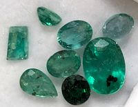 Close-up image showing eight faceted genuine emerald gemstones of varying shapes including oval, pear, rectangular, and round with natural inclusions.
