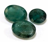 Three faceted oval-shaped emerald gemstones shown from an angled close-up, displaying natural green coloring and inclusions.