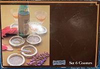 Coaster set box with photograph of silver plated coasters holding glasses and cigarette, showing item in use.