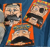 Three packages of self-adhesive moustaches displayed on blue cloth. Each package shows a different style of black moustache on illustrated faces.