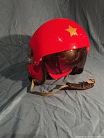 Left angled front view of the red fighter helmet showing the yellow star insignia, built-in clear and shaded visors, and chin strap with fabric padding.
