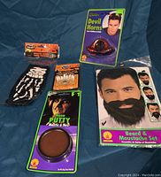 Overview of all items in the lot placed on blue fabric, showing packaging and items: skeleton gloves, flashing teeth, devil horns, nose putty, and beard and moustache set.