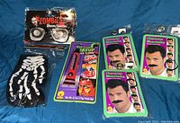 Full lot shown on blue fabric background including zombie glasses, skeleton gloves, red cream makeup, and three packaged character moustaches.
