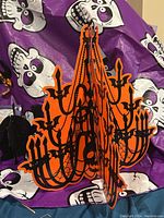Orange and black chandelier pumpkin hanging decoration in folded view on top of purple skull tablecloth.