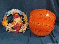 Three orange plastic bowls decorated with jack-o'-lantern faces stacked together next to a welcome door hanger with a scarecrow and fall leaves on a blue backdrop.