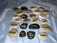 A top-down photo showing a collection of about 25 polished rocks with various inspirational words engraved in gold or black lettering. Rocks vary in color from beige to black and size from approximately 1 to 2.5 inches.
