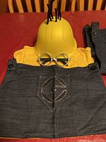 Full Minion costume components including yellow helmet, round glasses, black 'G' bib overall, and yellow T-shirt arranged on a red table