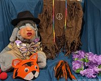 Wide view showing gray stuffed animal wearing kissy-face mask and butterfly mask with brown fringe vest and black hat behind; orange and black tiger hat and purple lei in front