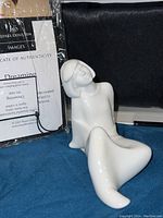 Photo showing white porcelain sculpture of a seated female figure with a certificate of authenticity behind it, certificate titled 'Dreaming' from Royal Doulton Images series, in original plastic packaging.