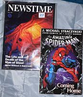 Two publications laid flat: On left is the May 1993 Newstime magazine with cover headline 'The Life and Death of the Man of Steel'. On right is The Amazing Spider-Man: Coming Home graphic novel cover featuring Spider-Man in red and blue suit.