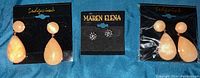 Photo showing three pairs of costume jewellery earrings placed on a blue fabric background. Two pairs are peach-colored teardrop earrings on cards labeled Sidgwick, and one pair of silver-toned stud earrings on a black card labeled Maren Elena.