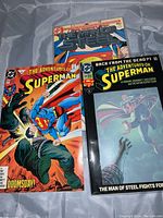 Photo showing three Superman comics with colorful covers: 'The Man of Steel', 'The Adventures of Superman' with Doomsday, and another 'Adventures of Superman' issue.