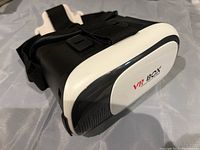 Front angled view of the VR Box headset showing the black and white color scheme with logo on front panel and adjustable straps.