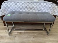 Front view of gray tufted upholstered bench with polished chrome metal legs in front of bed.