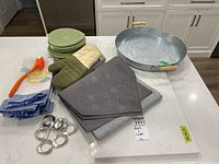 Photo showing quartz cutting board, metal tray, table runners, oven mitts, napkin rings, dish brush, tea towels, and four green plates on kitchen counter.