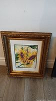 Full framed watercolor painting showing bright yellow sunflowers with green leaves in an ornate gold frame placed on floor against wall.
