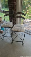 Two metal bar chairs with fabric seats, showing dark metal frame and script pattern upholstery, located by a window.