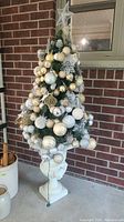 Full decorated artificial Christmas tree featuring assorted ornaments in gold, silver, white, cream with a star topper, placed on porch against brick wall.