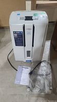 Front view of the white DeLonghi dehumidifier with instruction booklet and hoses.