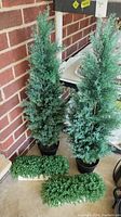 Two tall faux trees about 39 inches tall with dense green foliage and two small faux shrub-like plants placed on a surface against a brick wall.