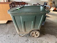 Side view of green plastic garbage bin on a metal dolly with two wheels, showing ribbed design and some dirt on the bin and wheels.