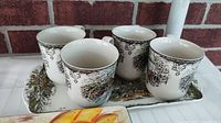 Four Johnson Bros mugs arranged in a row on a metal tray in front of a brick wall, showing the floral and countryside design pattern.