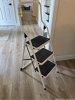 Side view of the open white folding metal step ladder showing three black steps with textured surface and handle.