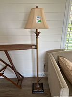 Full view of the floor lamp showing the metal base with floral decorative motifs and beige fabric shade in a living room setting.